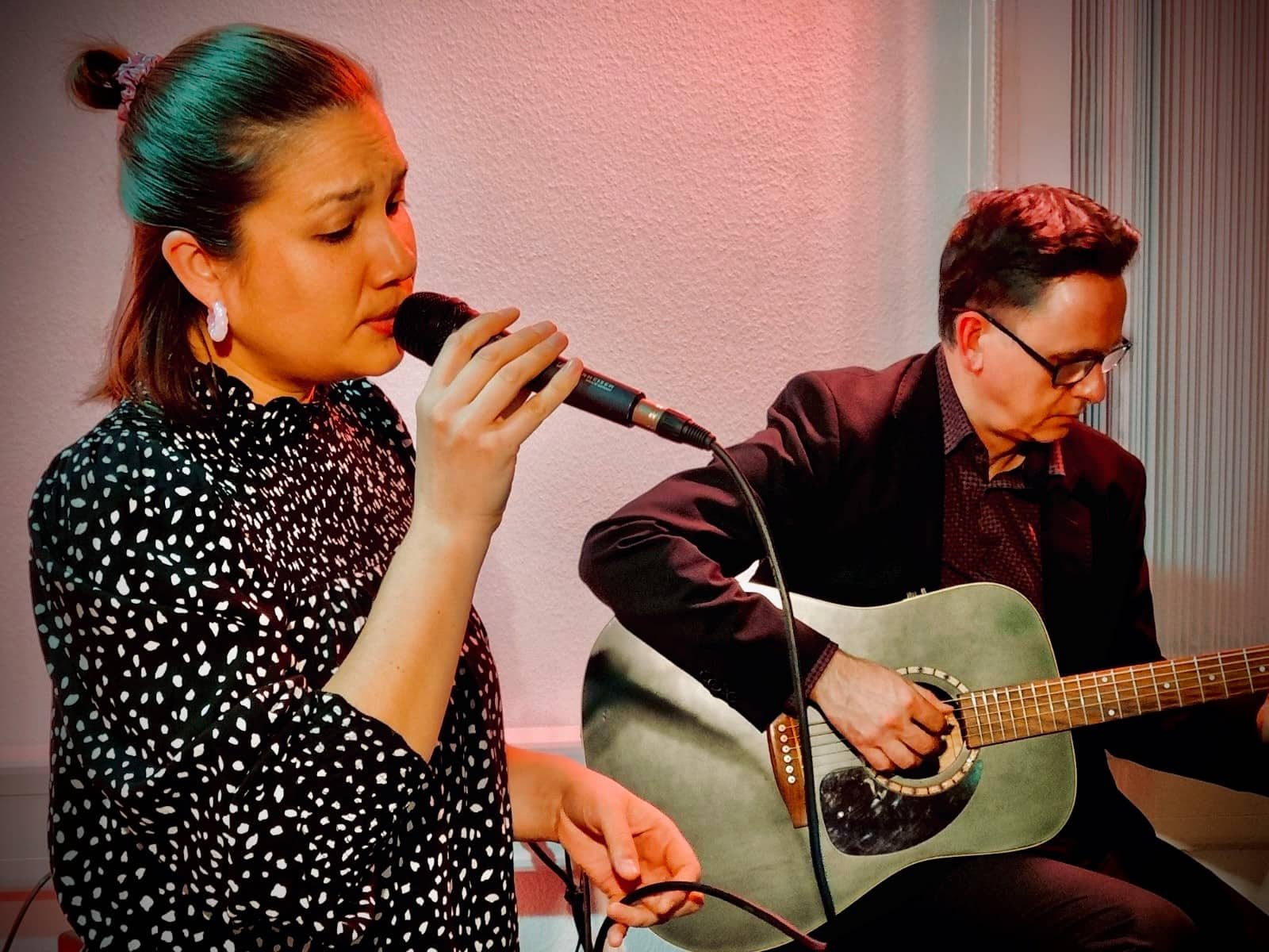 Frau mit schwarzem, weiß gepunktetem Oberteil hält Mikrofon, Mann spielt akustische Gitarre, beide sitzen vor weißer Wand mit Vorhang.